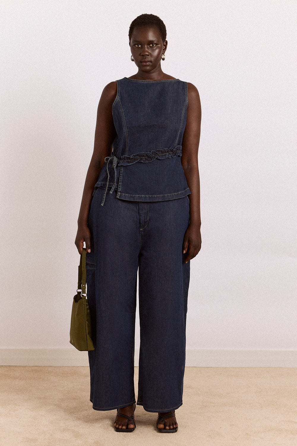 Woman wearing a navy sleeveless top and pants with a green bag against a white background