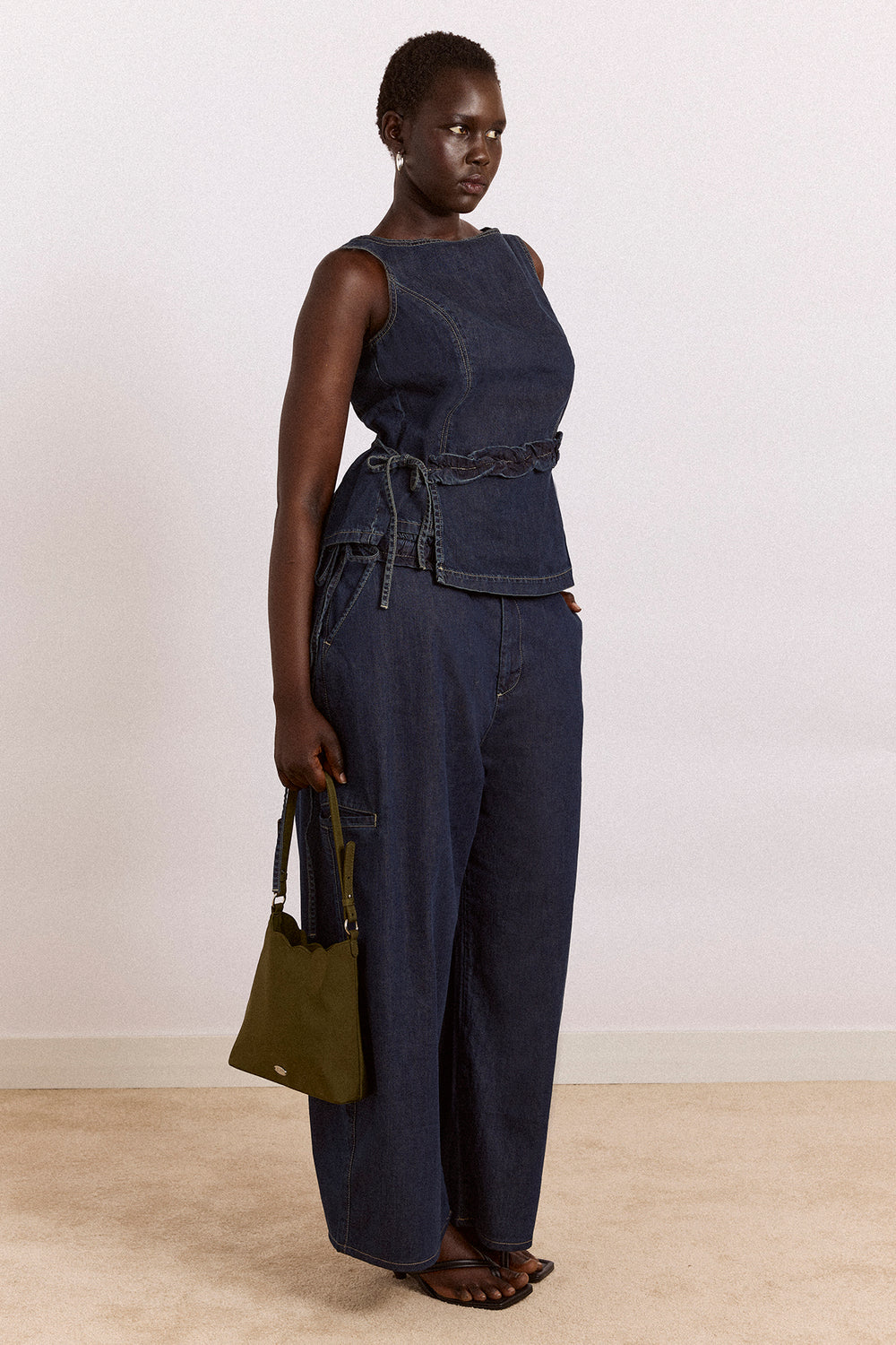 Woman wearing a dark blue sleeveless top and pants holding a green bag against a plain background